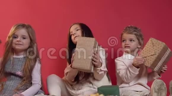 三个可爱的快乐小模特坐在红色背景的地板上玩着包装完好的礼物视频的预览图