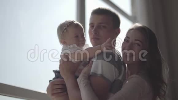 英俊的年轻人把小女孩抱在怀里在窗前特写妈妈站在她丈夫旁边视频的预览图