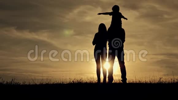 日落时分幸福家庭的剪影孩子坐在父亲的脖子上从后面看视频的预览图
