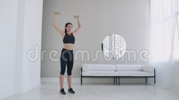 一个年轻的黑发女人用手缓慢地举起哑铃为一个美丽的身体做家庭锻炼视频的预览图