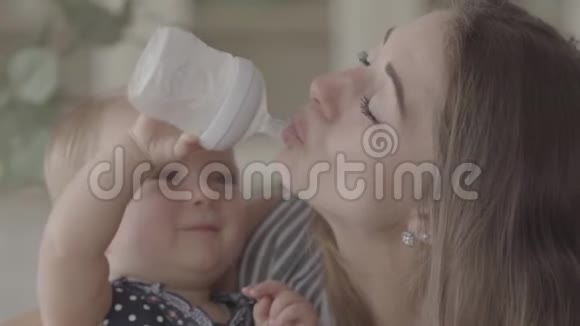 美丽微笑的女婴试图从厨房的婴儿瓶里给她的母亲喝水幸福的概念视频的预览图