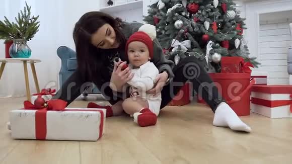可爱的婴儿戴着筒状帽子坐在圣诞树附近的地板上他可爱的母亲想修理红球视频的预览图