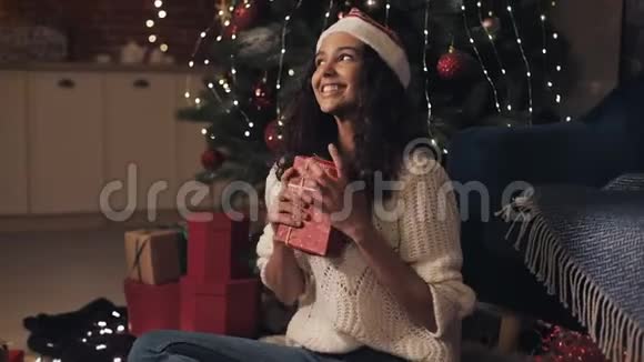 圣诞帽里的年轻美丽的穆拉托女孩坐在装饰的圣诞树拥抱着红色的礼物盒里看上去很快乐视频的预览图