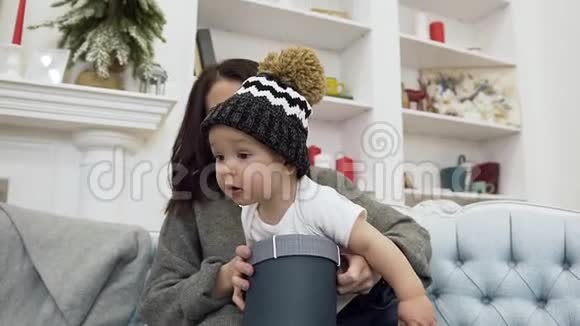 可爱的蹒跚学步的孩子啃着小纸盒他可爱的年轻母亲和他一起坐在舒适的沙发上视频的预览图