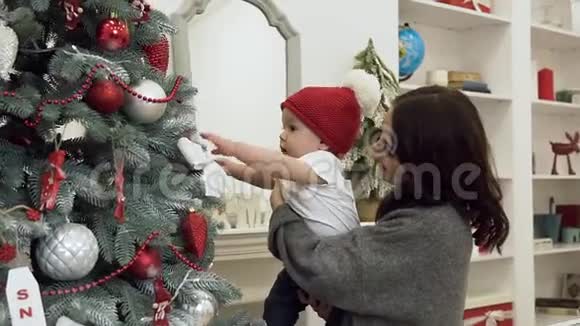 漂亮的微笑妈妈和可爱的婴儿站在装饰好的圣诞树孩子试图得到圣诞树视频的预览图