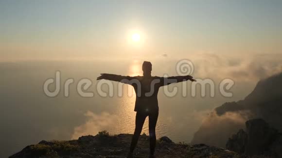 一位年轻女子伸出双臂从一座高山望着海面上美丽的落日视频的预览图