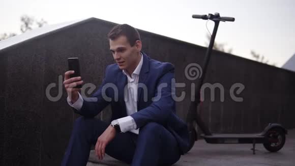 漂亮的商人坐在楼梯上在外面的智能手机上做视频穿着优雅蓝色西装的男人视频的预览图
