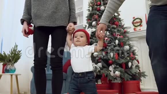 令人难以置信的可爱的蹒跚学步的孩子的慢动作他双手牵着父母走在圣诞树附近视频的预览图