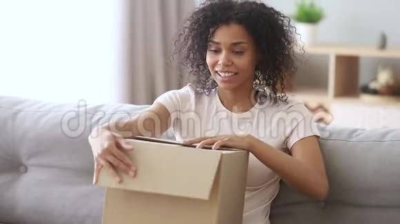 快乐的非洲女人顾客打开纸板箱坐在沙发上视频的预览图