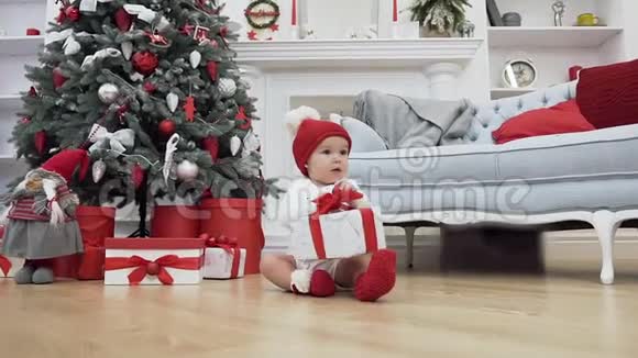 一个穿着圣诞衣服的可爱的小宝宝坐在地板上拿着礼物在附近的圣诞照片视频的预览图