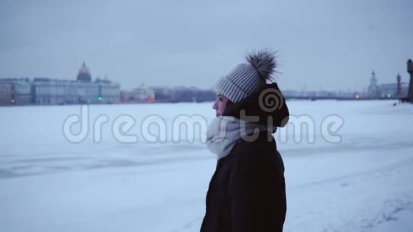 穿着温暖夹克和帽子的年轻女孩在寒冷中年轻的女人在雪堆和雪城的背景下冬季视频的预览图