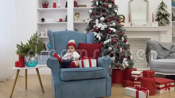 美丽可爱的蹒跚学步的孩子坐在蓝色的大扶手椅上枕在节日装饰的圣诞礼物中视频的预览图