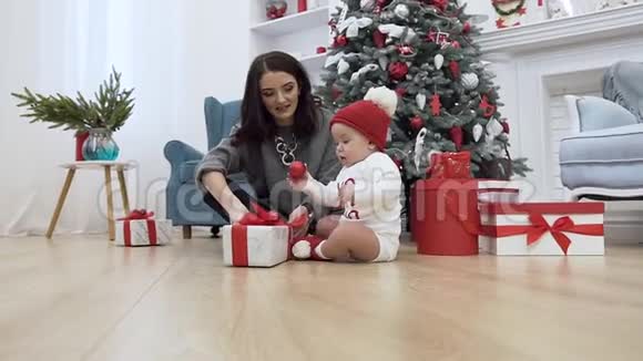 漂亮的母亲坐在圣诞树附近的地板上她可爱的婴儿试图接住红色的球视频的预览图