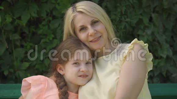 一幅成熟的金发母亲戴着眼镜小女儿坐在长凳上看着摄像机的画像视频的预览图