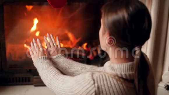 穿着毛衣的年轻女子在屋里燃烧的壁炉旁暖和搓手的4k镜头视频的预览图