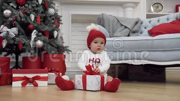 戴着圣诞帽的可爱宝宝坐在仙女圣诞树旁试着打开圣诞礼物视频的预览图