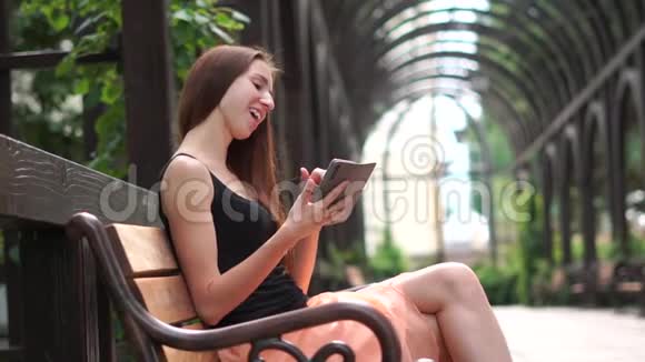 女孩高兴地在智能手机上读到一些东西坐在长凳上的公园里视频的预览图