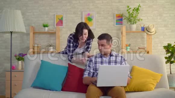 年轻积极的妻子高兴地跳舞她怀孕了悲伤的丈夫震惊地坐在沙发上的笔记本电脑前视频的预览图
