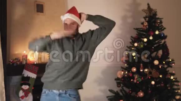 快乐的留胡子的人在圣诞树附近跳舞戴着圣诞老人帽子的快乐英俊男人以滑稽的方式移动他的身体很漂亮视频的预览图