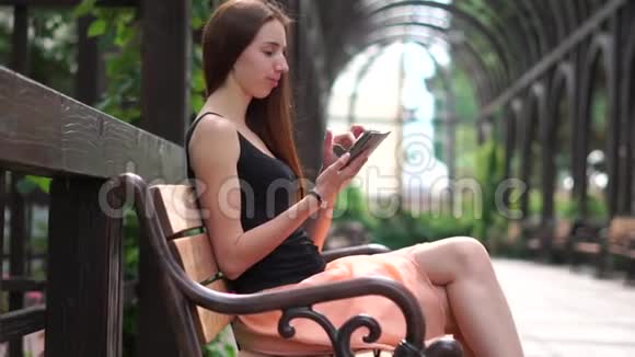 女孩坐在长凳上在公园的智能手机上打印一条信息慢动作视频的预览图