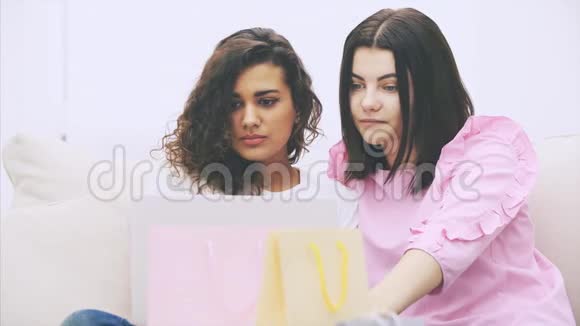两个快乐的女朋友坐在沙发上在家里拿着笔记本电脑选择和讨论网上买什么模糊的视频的预览图