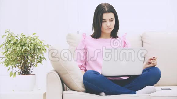 可爱的白种人黑发女孩坐在沙发上拿着笔记本电脑上网扔手视频的预览图