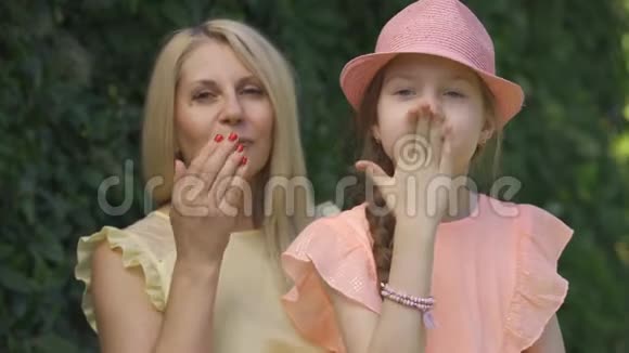 金发母亲和她的小女儿坐在夏天的公园里一边看着镜头一边送空气亲吻的肖像视频的预览图