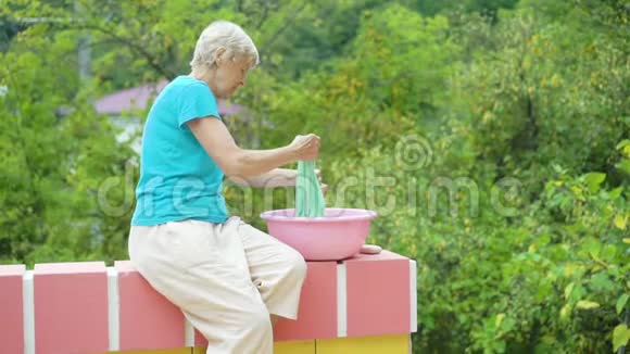 一位老妇人穿着粉色塑料盆在大自然绿色的背景下洗衣服视频的预览图