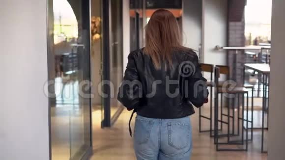 一个年轻漂亮的白种人女孩穿着黑色皮夹克和牛仔裤走在购物中心转过身笑了笑视频的预览图