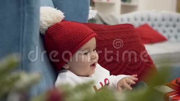 把戴着红帽子戴着白头巾坐在一张带枕头的大椅子上拿着礼物玩的可爱的幼儿合上视频的预览图