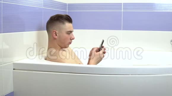 一个年轻人躺在浴室里手里拿着手机一个男人在洗澡和聊天中放松视频的预览图
