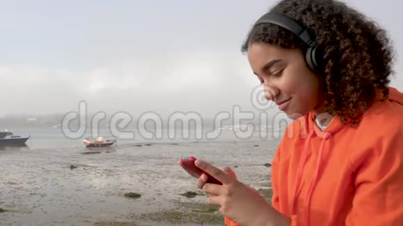美丽的混血非裔美国少女使用智能手机和耳机在海边喝咖啡视频的预览图