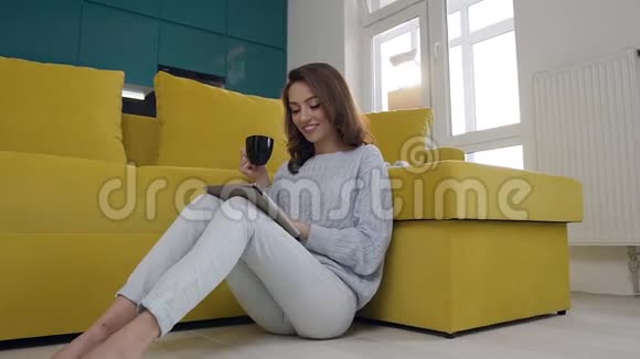 美丽的微笑快乐的女士坐在沙发附近喝着咖啡在厨房背景上使用她的平板电脑视频的预览图