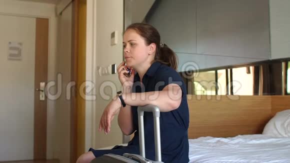 一个女人坐在酒店房间里躺在一个手提箱旁边的床上打电话视频的预览图