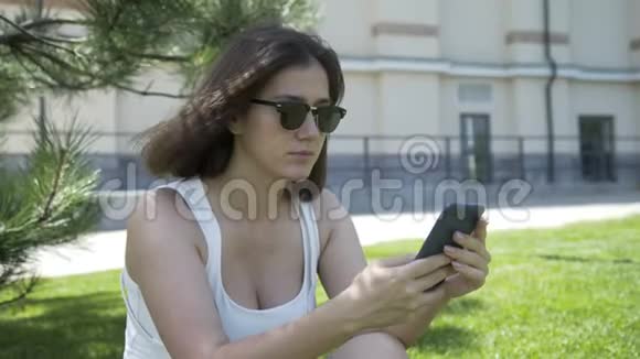 穿着白色T恤和太阳镜的年轻美女在办公大楼背景上的智能手机上发短信的画像视频的预览图
