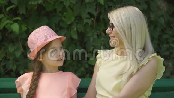 可爱成熟的戴着眼镜的金发母亲和她的小女儿在夏天的公园里坐在长凳上聊天视频的预览图