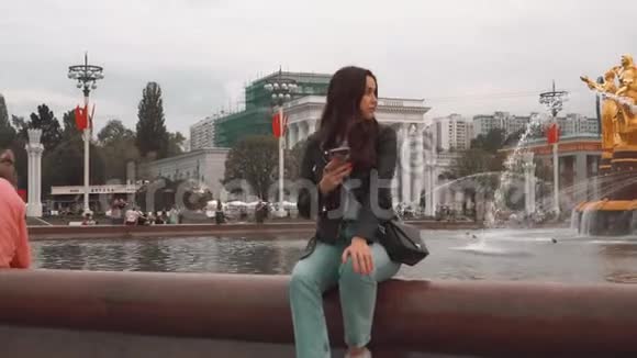 穿着夹克和牛仔裤的漂亮女孩坐在智能手机喷泉附近视频的预览图