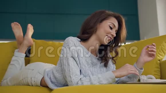 愉悦快乐的年轻女性卷发躺在舒适的沙发上使用平板电脑视频的预览图