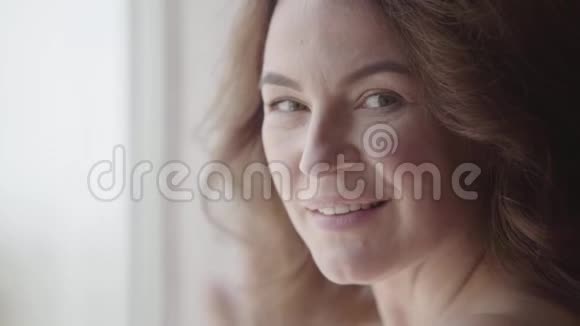 一个相当成熟的女人看镜头的特写肖像阳光从窗户照在她身上快乐的白种人女人视频的预览图