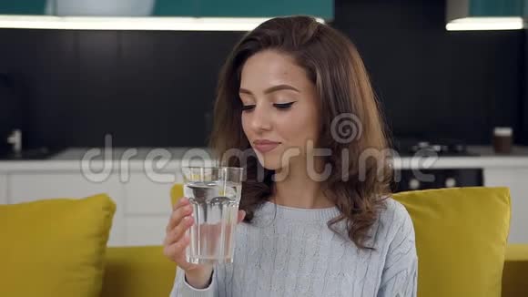 欢乐灿烂的年轻女子坐在浅绿色的沙发上在厨房里喝着一杯干净的水视频的预览图