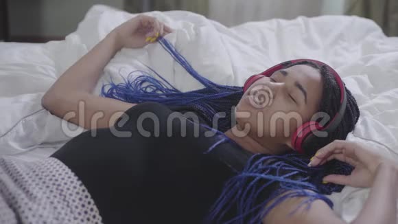 可爱的非洲裔美国女孩戴着耳机躺在白色床上的肖像眼睛紧闭头部移动年轻女子视频的预览图