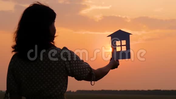 一个穿着裙子的女孩拿着纸房子的剪影夜晚美丽的日落家庭幸福和舒适视频的预览图