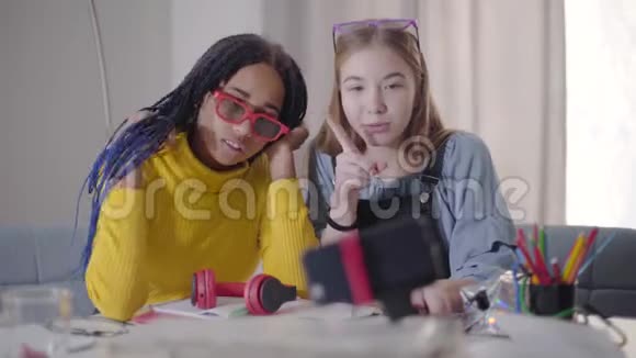 开朗的白种人金发女孩和她的非洲裔美国朋友扎着辫子在智能手机上的自拍相机上交谈视频的预览图