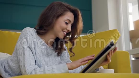 迷人的年轻微笑的女人棕色波浪发躺在舒适的沙发上用她的平板电脑视频的预览图