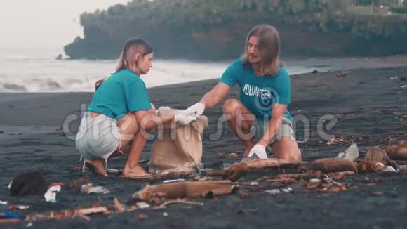 两名志愿者穿着蓝色t恤在海滩上清理垃圾把垃圾折叠在生态袋里视频的预览图