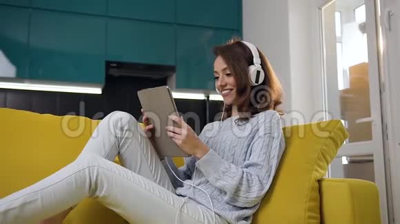 一个30岁的迷人的快乐女孩带着波浪头发坐在舒适的沙发上手提电脑听着音乐镜头视频的预览图