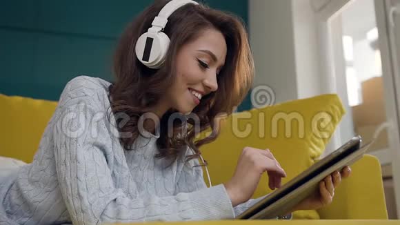 30岁戴着耳机听音乐用ipad躺在浅绿色沙发上的美女笑颜特写视频的预览图