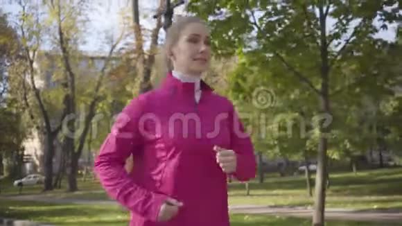 一个穿着粉红色运动服和耳机在秋天公园跑步的微笑白种人女孩的特写女运动员视频的预览图