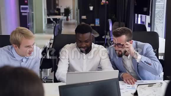 三位自信的年轻男性商业伙伴在电脑前合作讨论他们的商业项目视频的预览图
