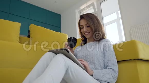 漂亮可爱的微笑女人坐在厨房沙发附近的地板上喝着芬芳的咖啡用她的方式视频的预览图